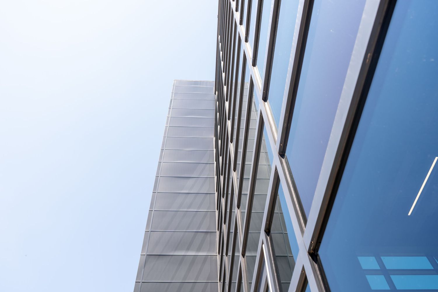 Modern glass office building facade with diagonal lines and reflective windows photographed from below against blue sky.