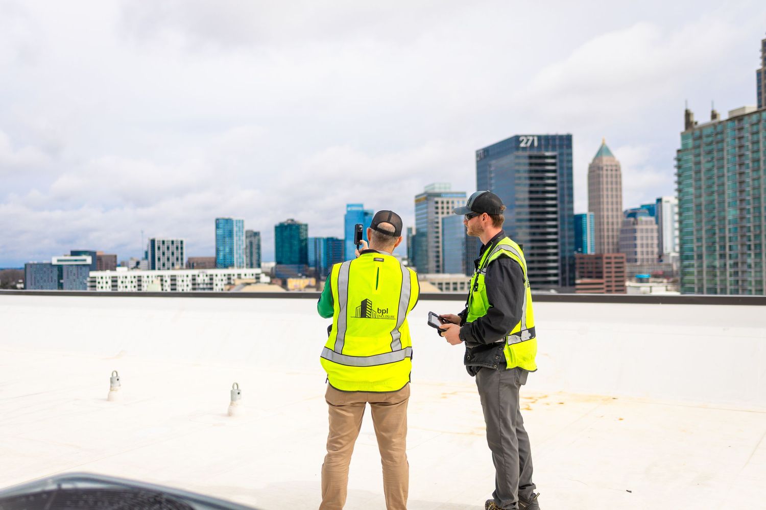Construction workers in safety vests review plans on a rooftop with city skyline in background.