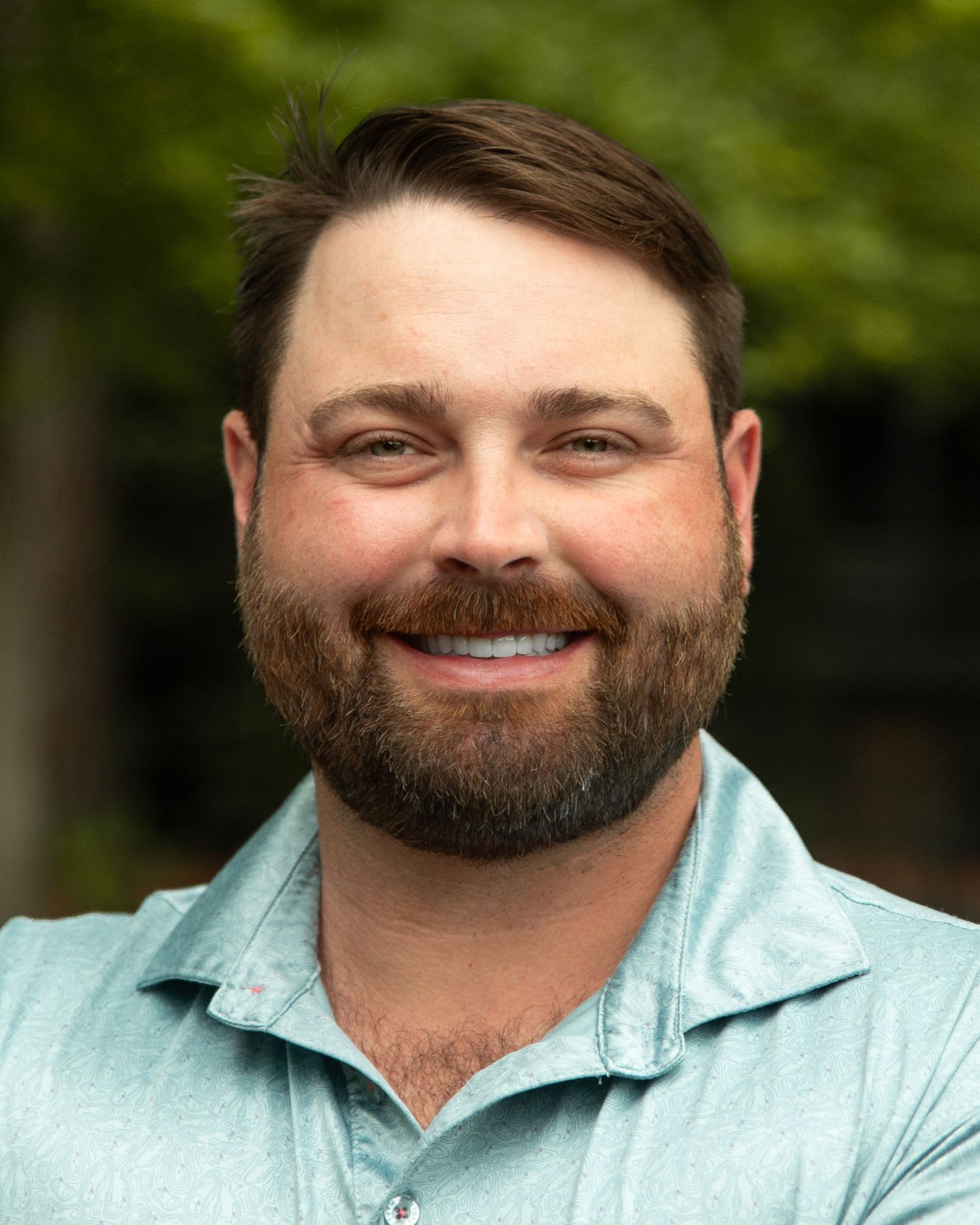 Smiling man with brown beard wearing a light blue polo shirt outdoors with green trees in background.