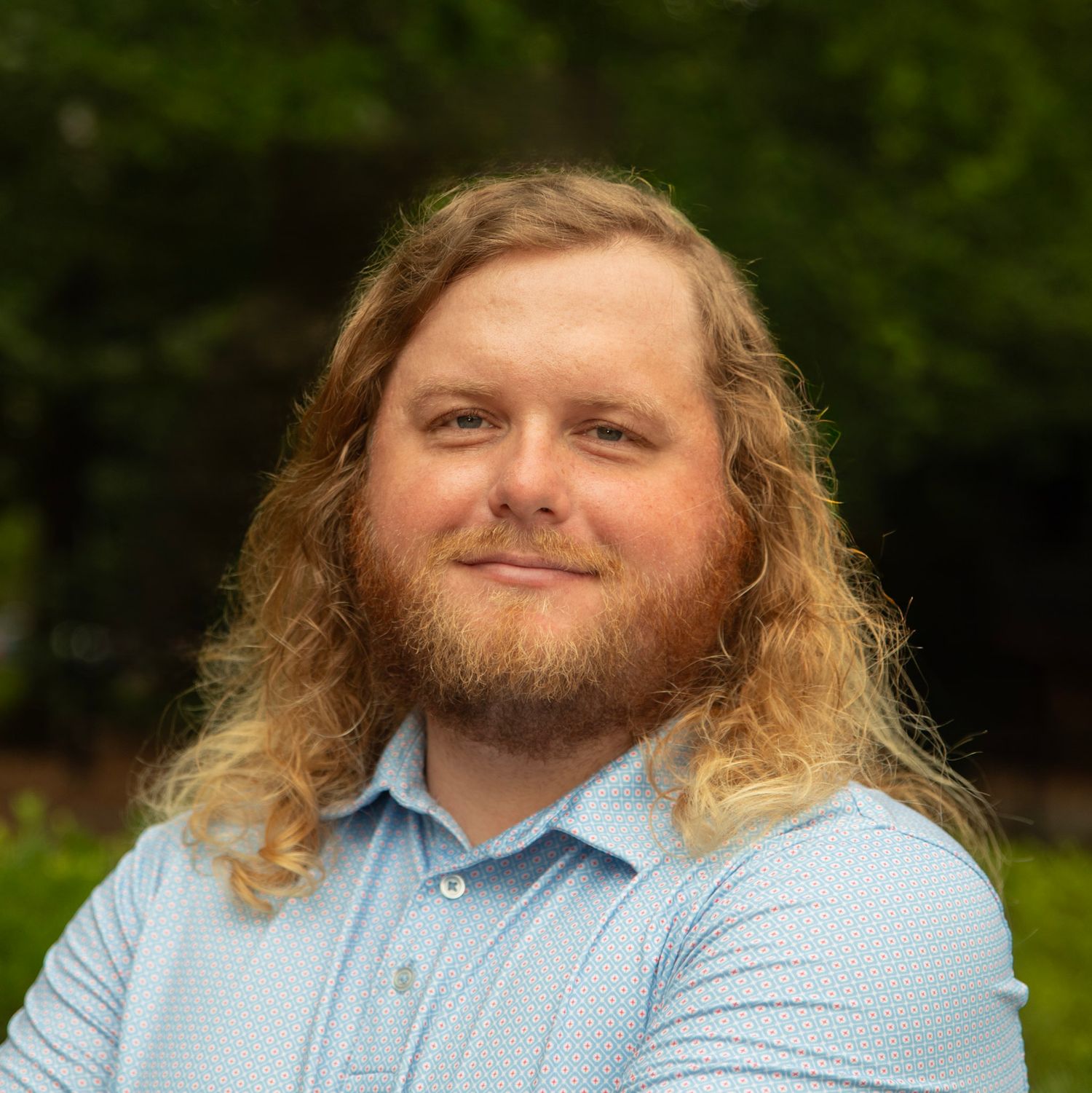 Smiling young man with long curly blonde hair and beard wearing a light blue shirt outdoors.