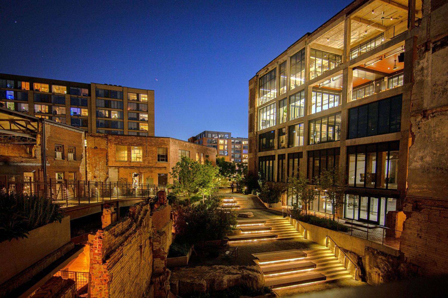 Illuminated urban courtyard with modern glass buildings and lit stairway at twilight in a city setting.