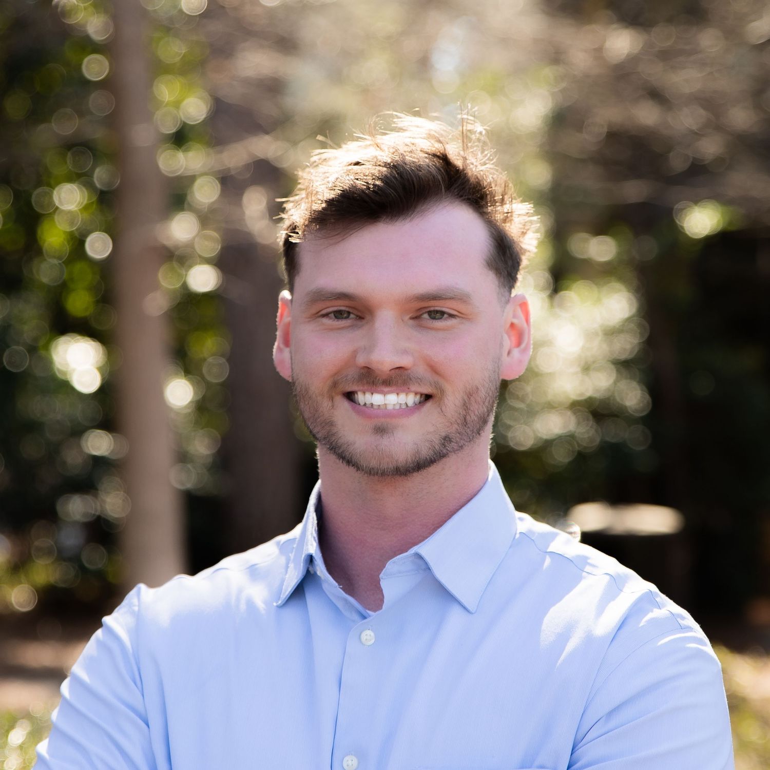 Person wearing a light blue button-down shirt photographed outdoors with blurred natural background in sunlight.