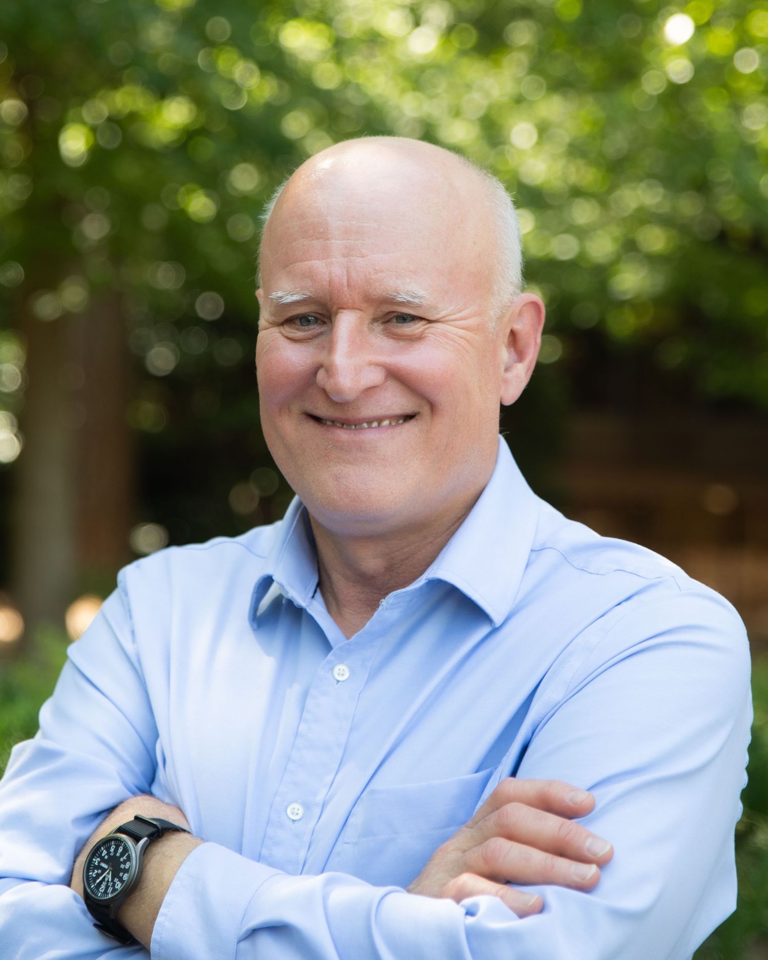 Smiling bald man with crossed arms wearing a light blue shirt and watch, standing outdoors with green trees.