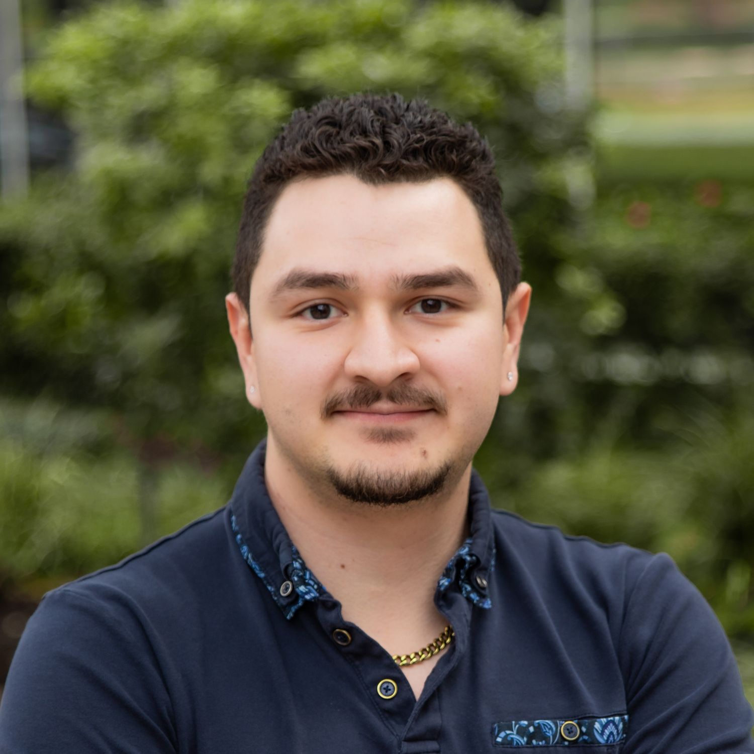 Professional headshot of someone in a navy polo shirt with embroidered details against a blurred green outdoor background.