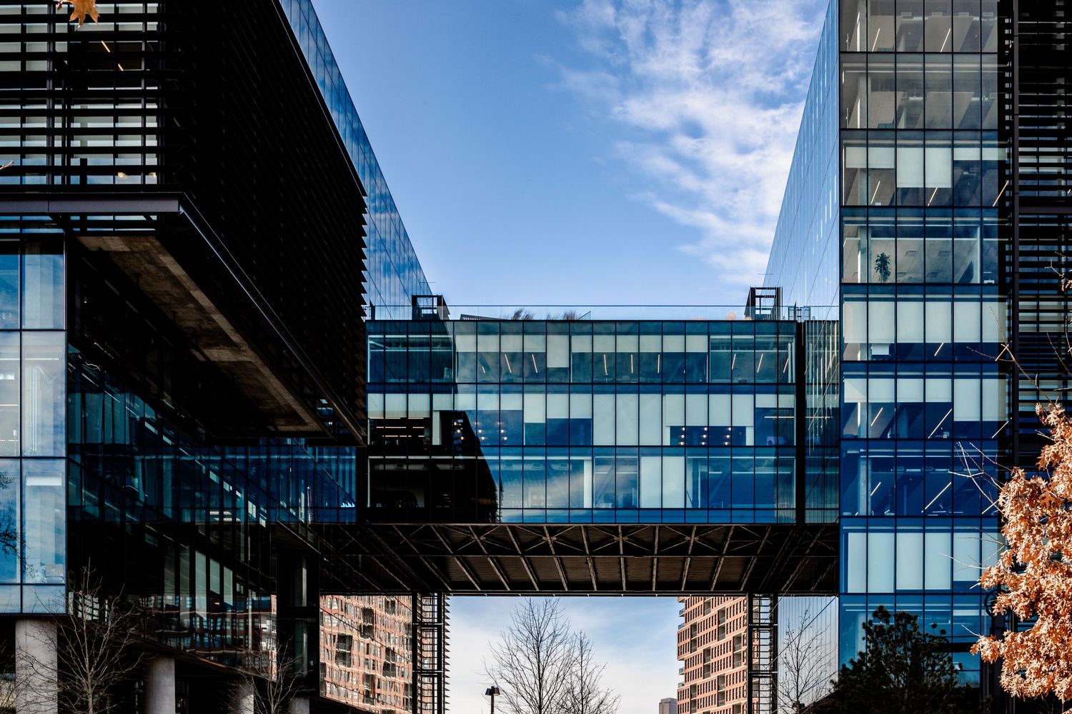 Modern glass office building with a bridge connecting two towers against a blue sky with white clouds.
