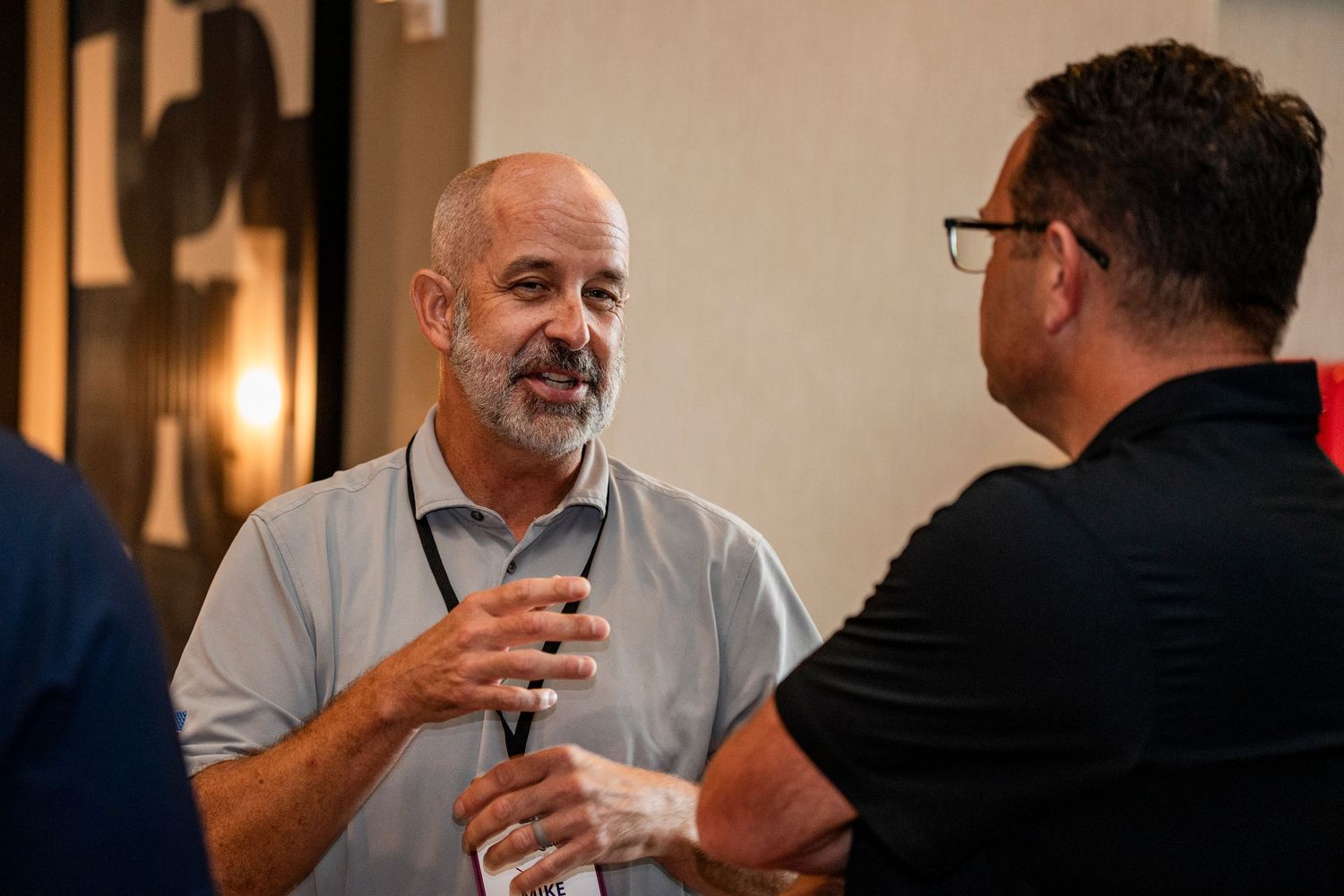 Two men engaged in animated conversation at an indoor networking event, one wearing a light gray shirt.