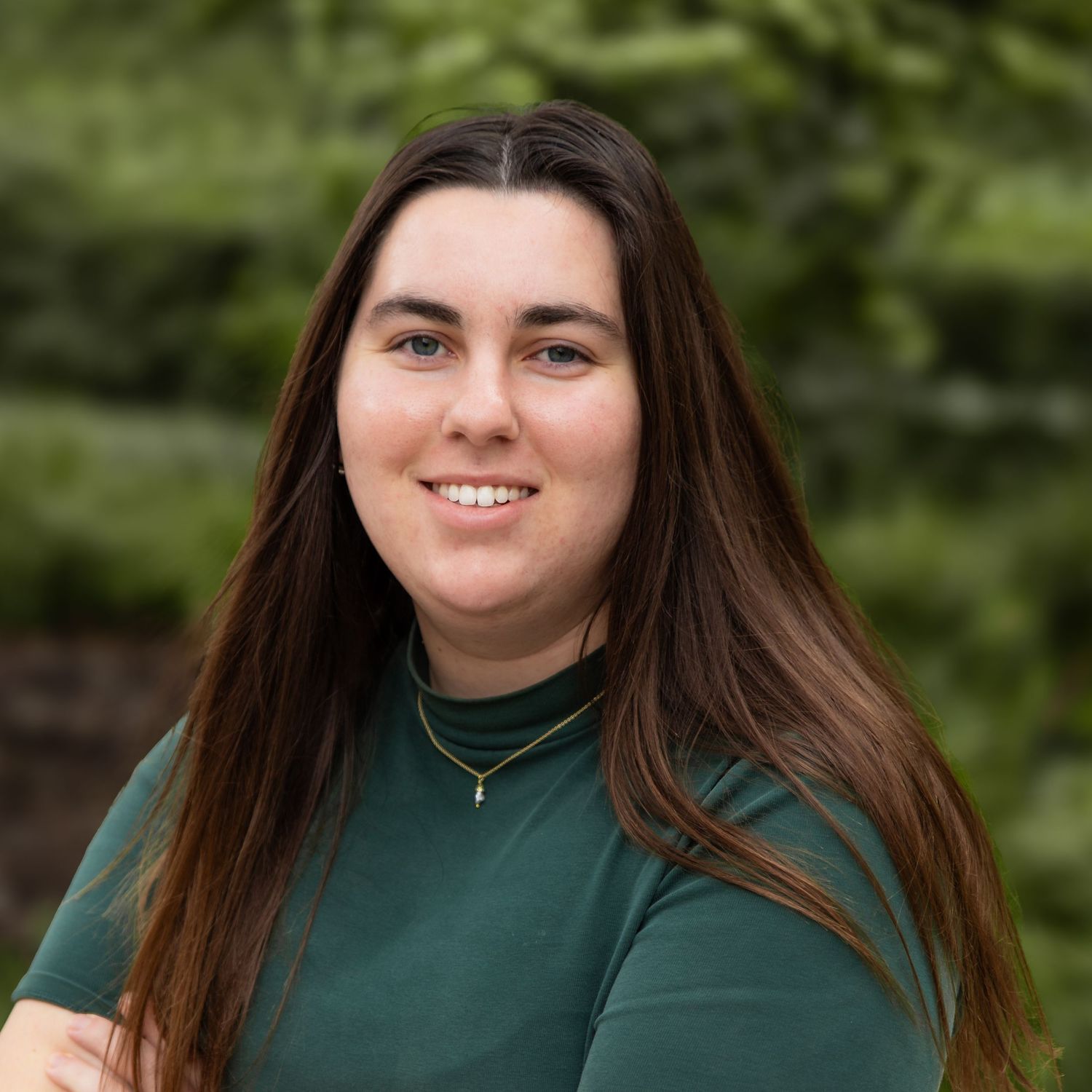 Person in dark green turtleneck sweater poses outdoors against natural foliage background.