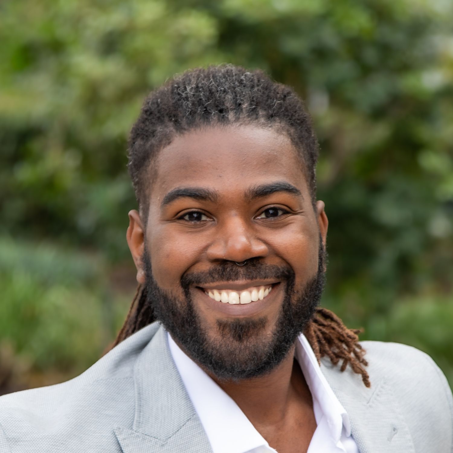 Professional headshot of a person with a beard and dreadlocks wearing a light gray blazer and white shirt, smiling warmly.