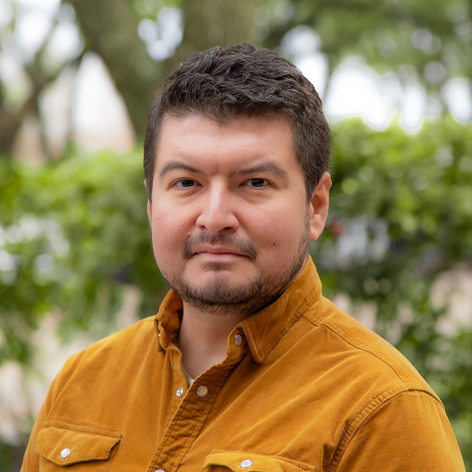 Professional headshot of someone wearing a mustard yellow button-up jacket outdoors with green foliage in background.