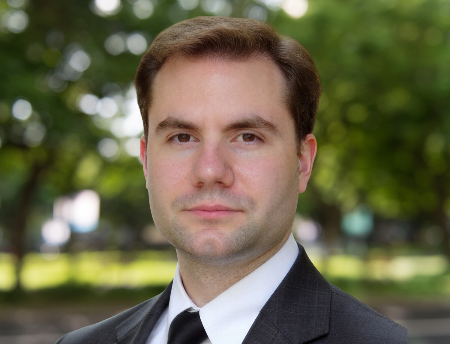 Professional headshot of a young businessman in a dark suit and tie outdoors with green trees in background.