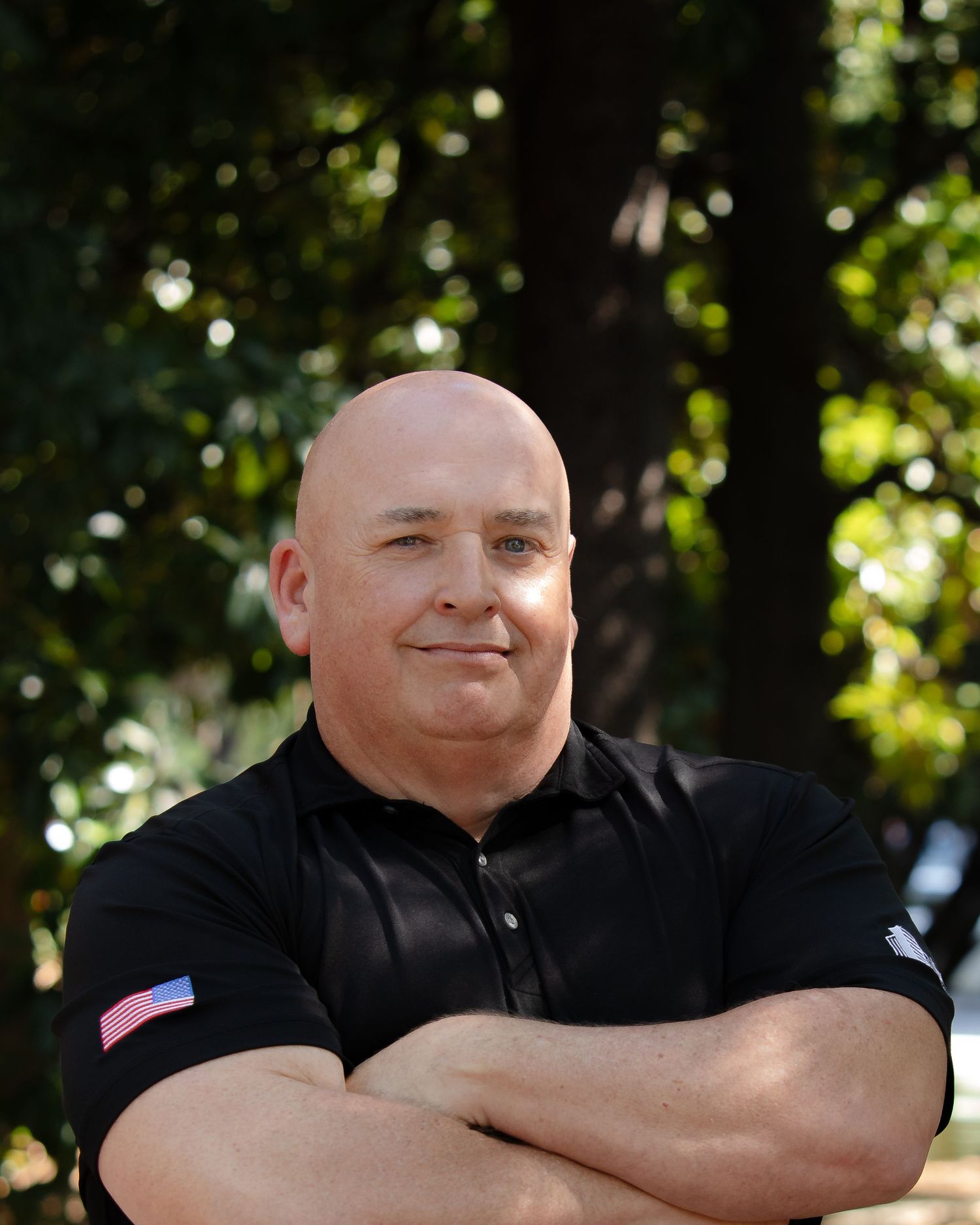 A person in a black shirt smiles while posing outdoors with trees and foliage in the background.