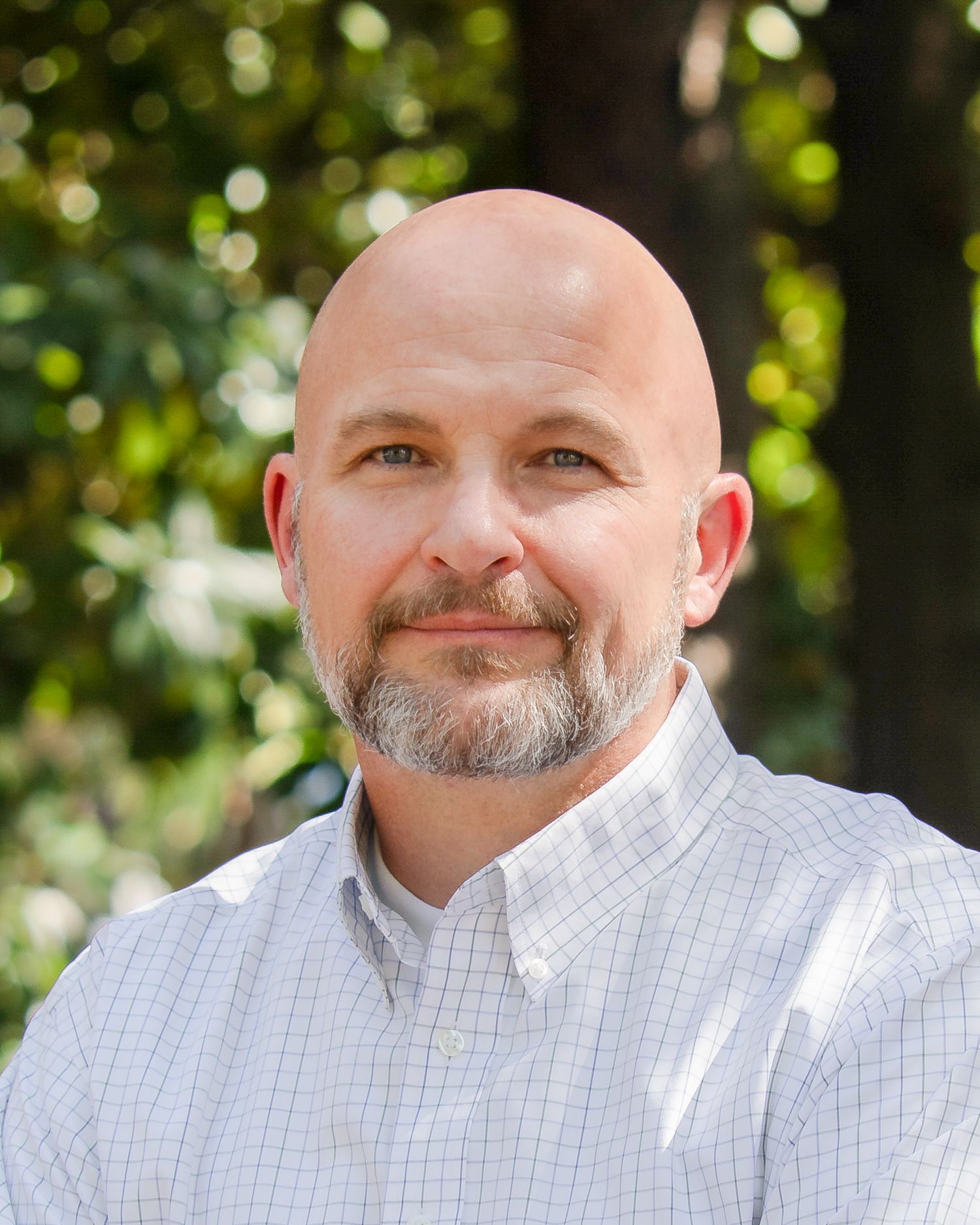 A person with a gray beard wearing a white button-down shirt smiles while standing outdoors with bokeh background.