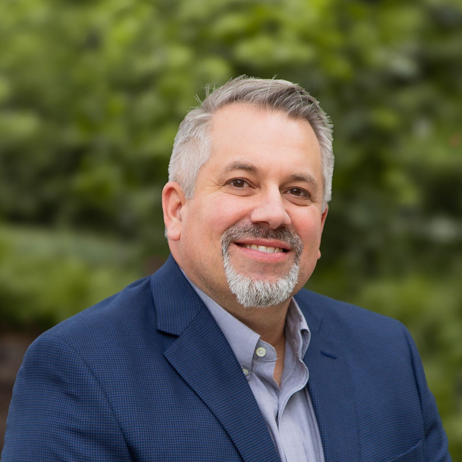 Professional headshot of a person in a navy suit jacket and gray shirt against a blurred outdoor background.