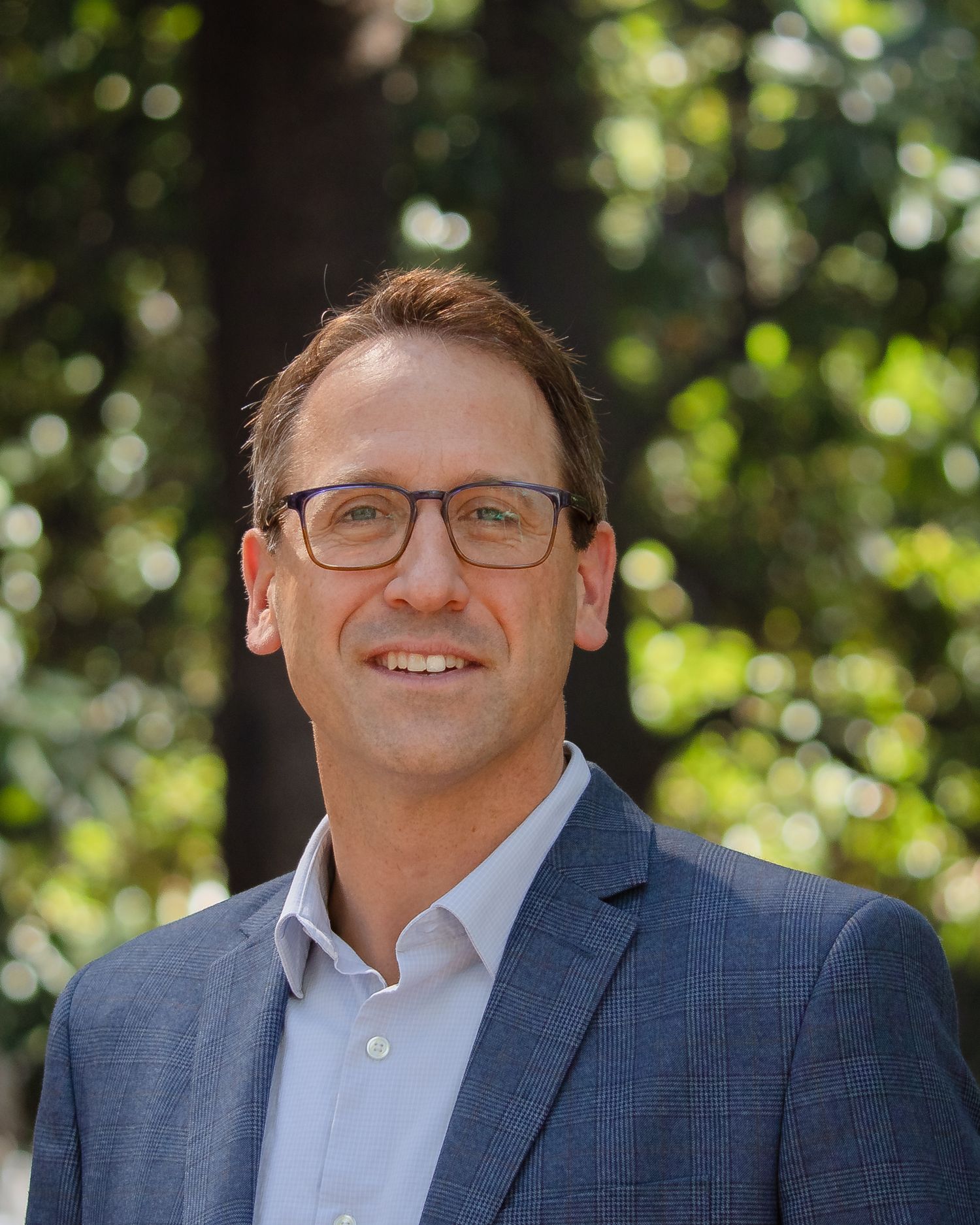 Professional headshot of someone wearing glasses and a navy suit jacket against a blurred natural outdoor background.