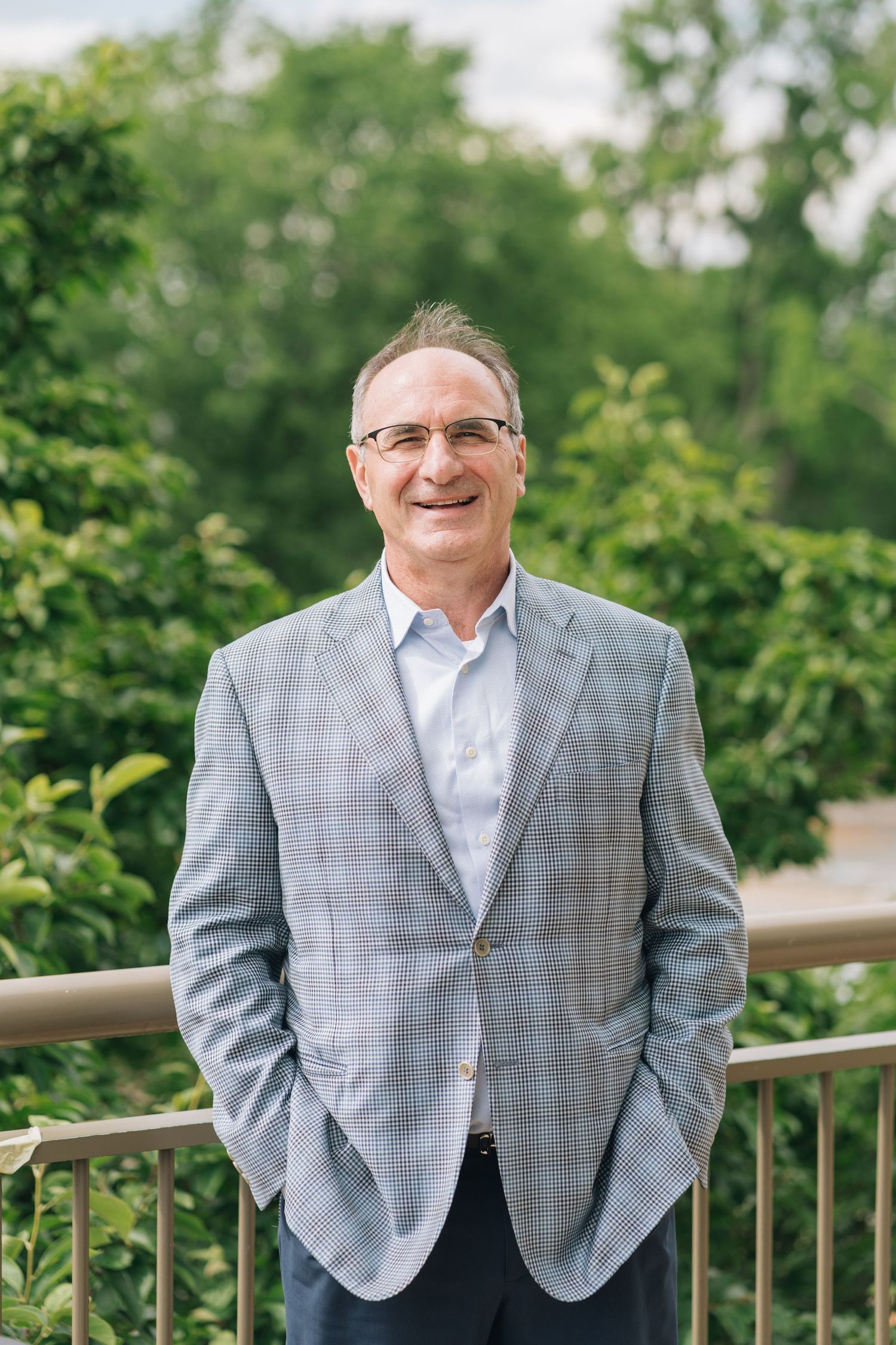 Professional wearing a gray plaid suit jacket stands outdoors against a natural green background.