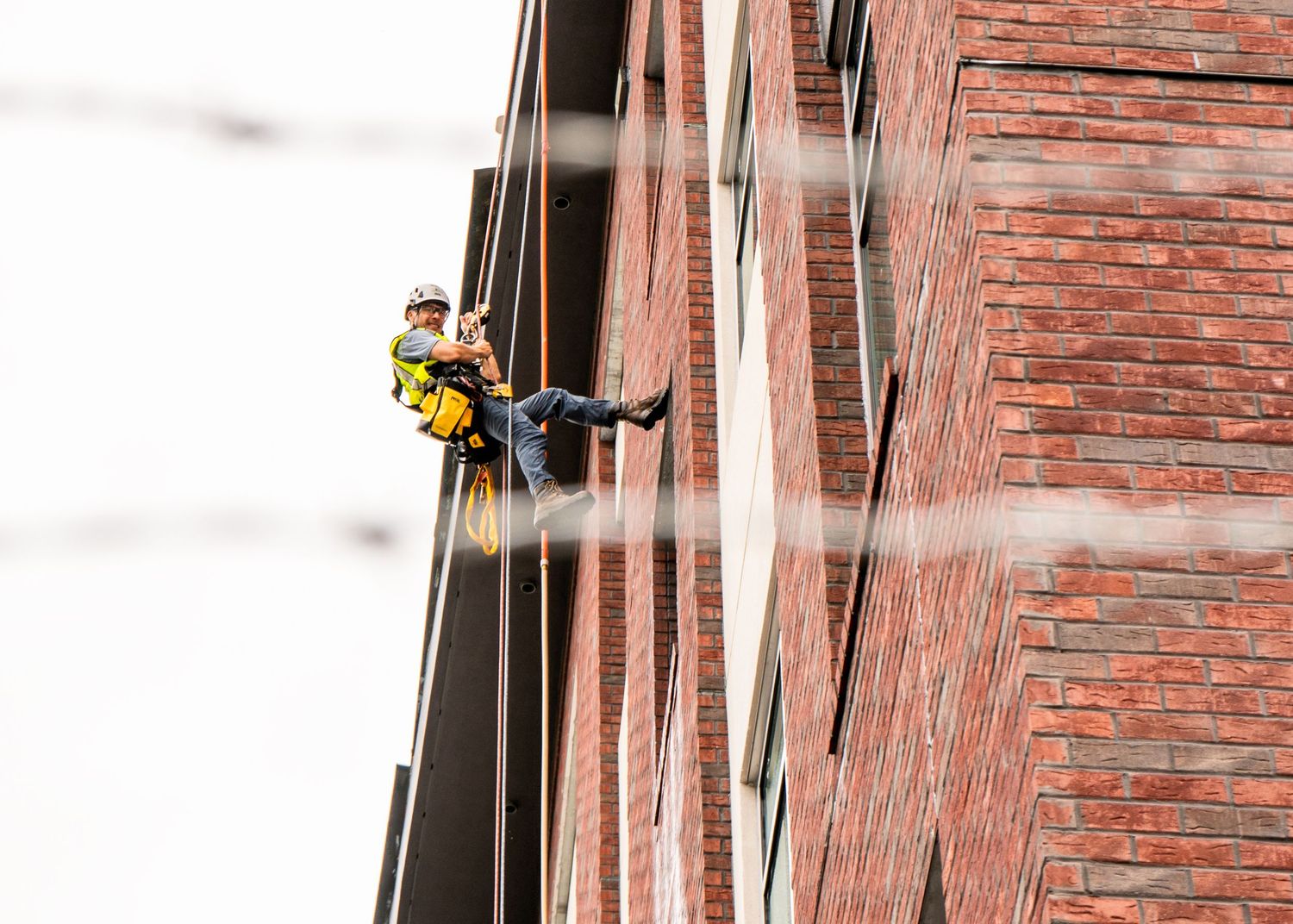 Professional window washer in safety harness rappels down brick building exterior while cleaning windows.