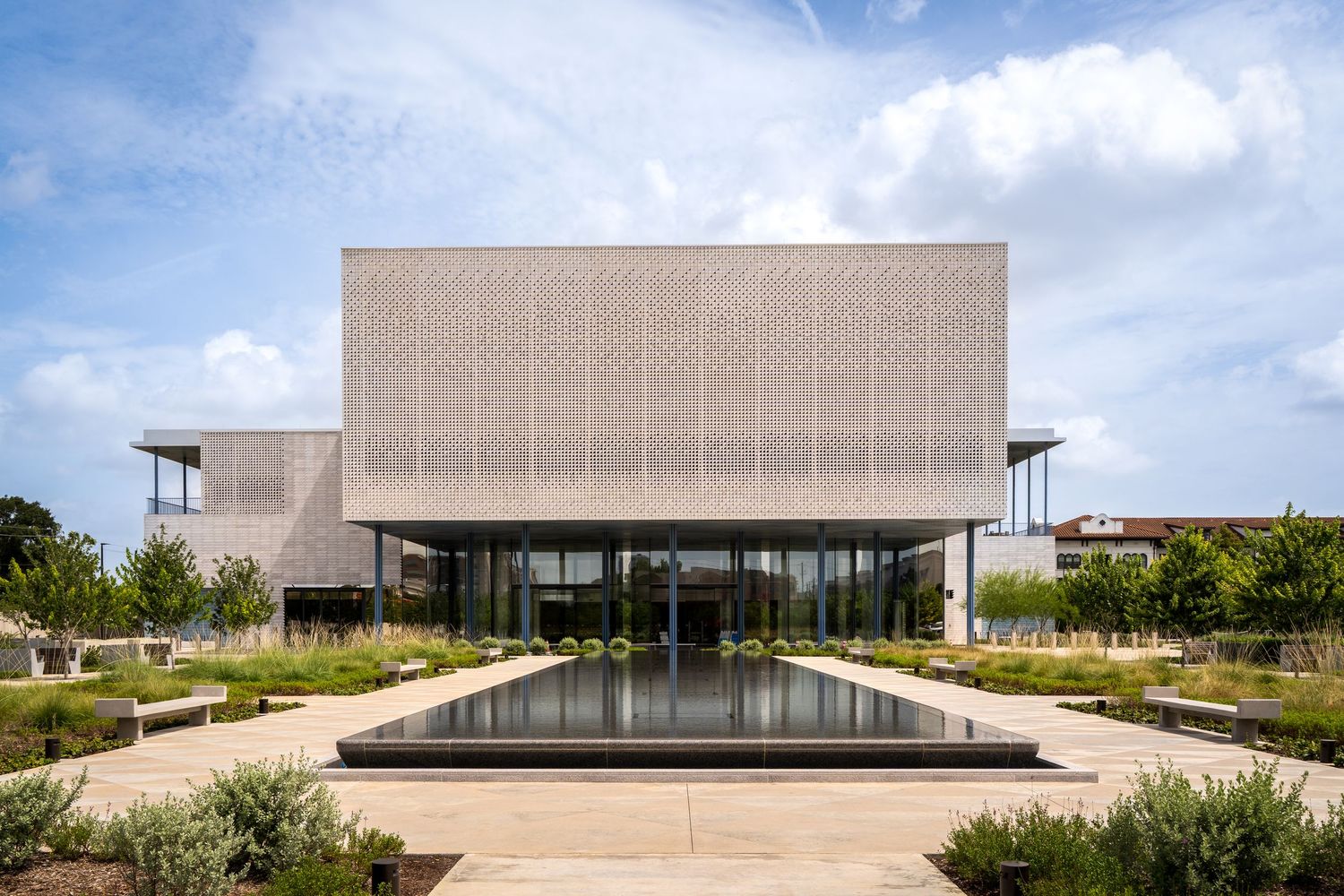 Modern museum building with decorative perforated facade, reflective pool, and landscaped gardens under a partly cloudy sky.