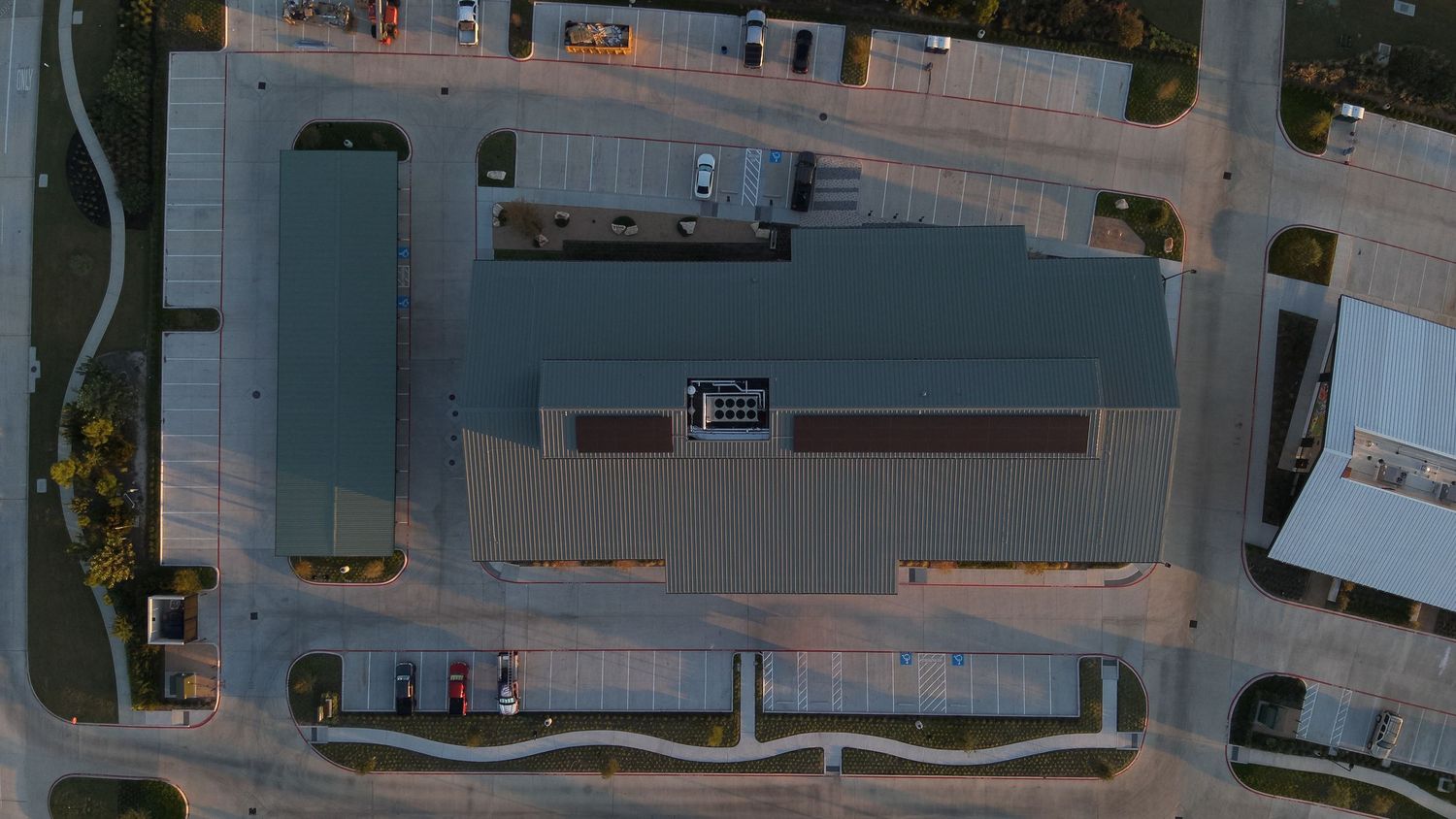 Aerial drone view of a large commercial building with gray metal roof surrounded by parking lots at sunset.
