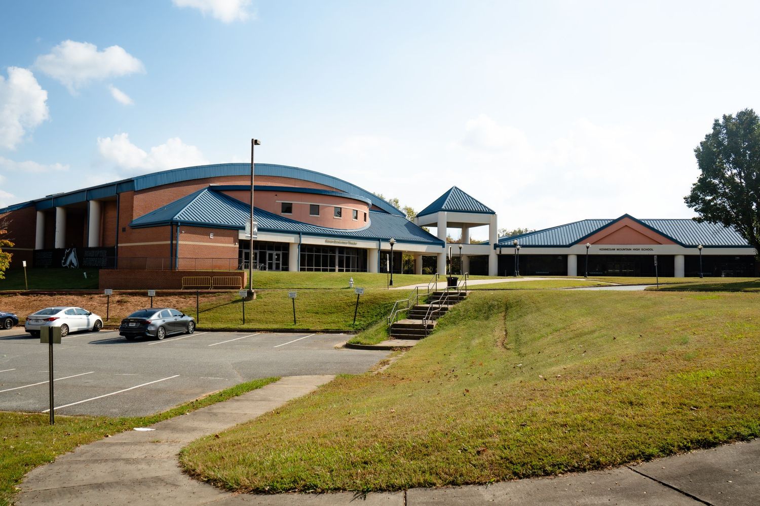 A panoramic view of modern school buildings and parking lots with grassy hills under a partly cloudy sky.