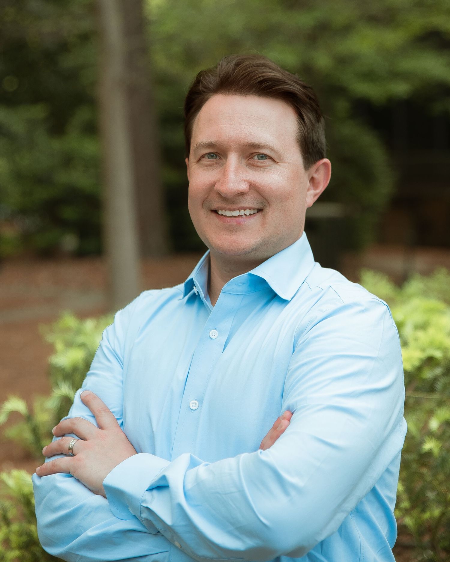 Smiling man in light blue shirt with arms crossed poses outdoors among lush green trees and shrubs.