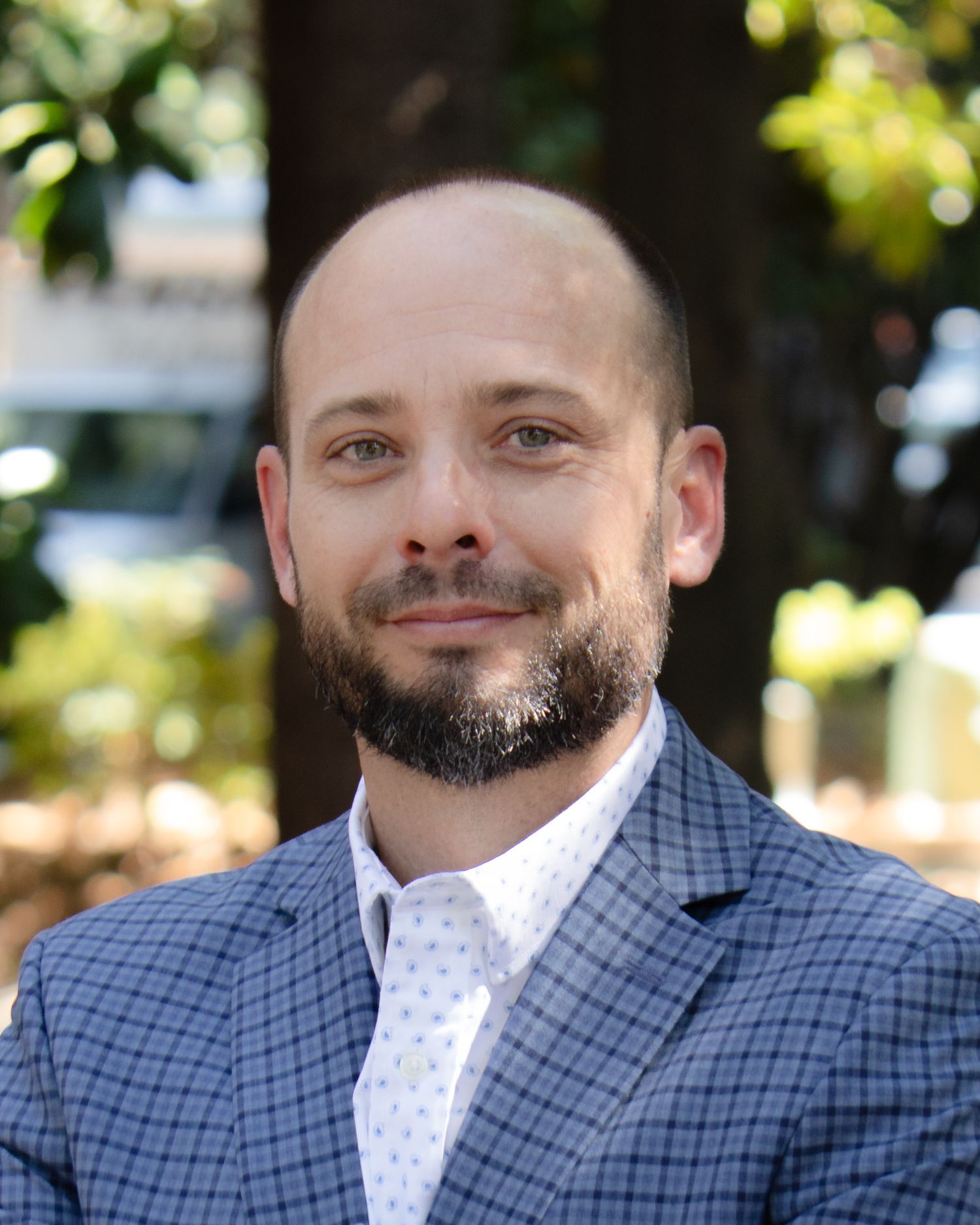 A man wearing a blue plaid blazer and white polka dot shirt smiles while looking to the side outdoors.