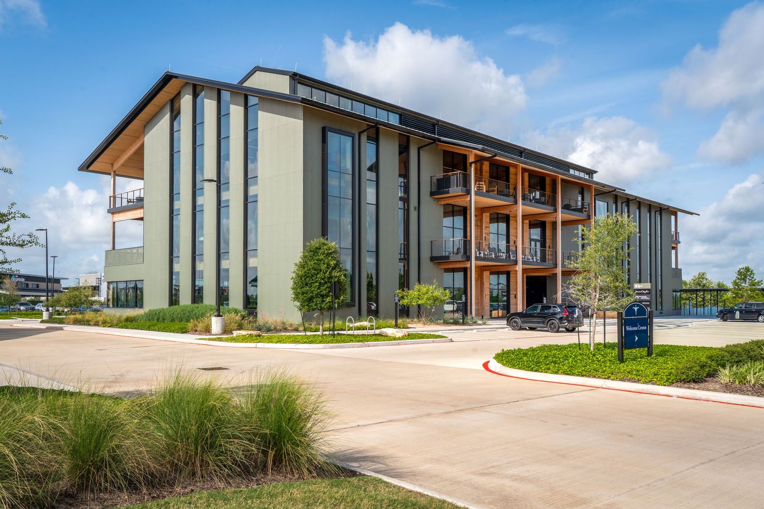 Modern three-story commercial office building with wood accents, large windows, and landscaped grounds under a partly cloudy sky.