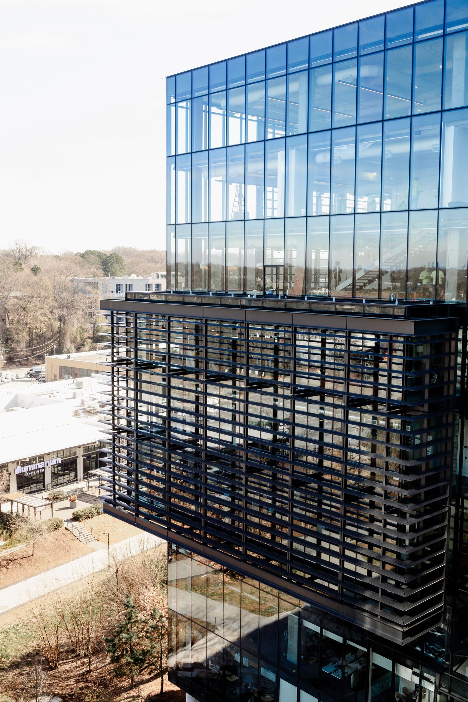 Modern glass office building exterior with black horizontal sunshades and reflective windows against a bright blue sky.