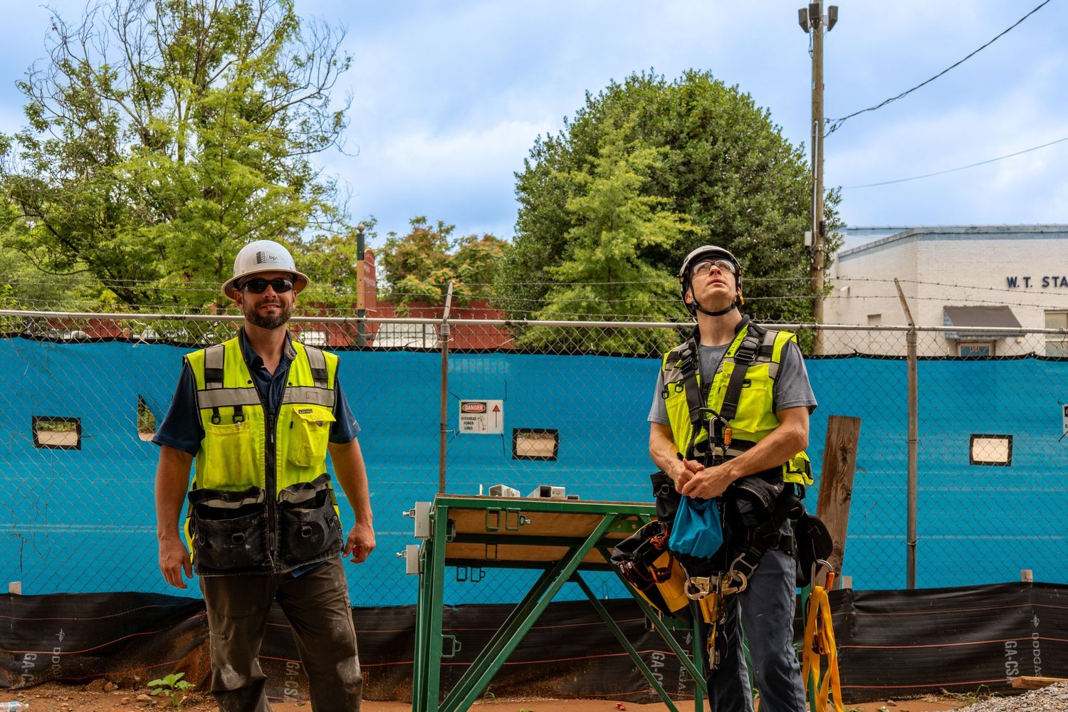 Two construction workers in safety vests and hard hats stand at a job site with blue fencing and trees in background.