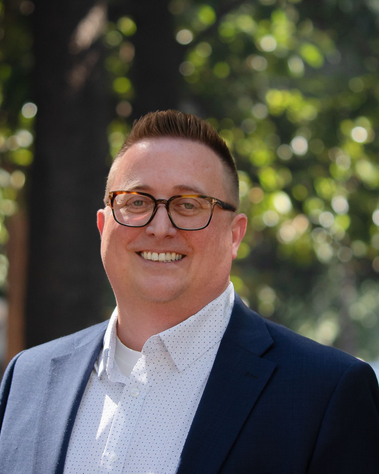 Professional headshot portrait of someone in glasses and navy suit with white polka dot shirt smiling outdoors.