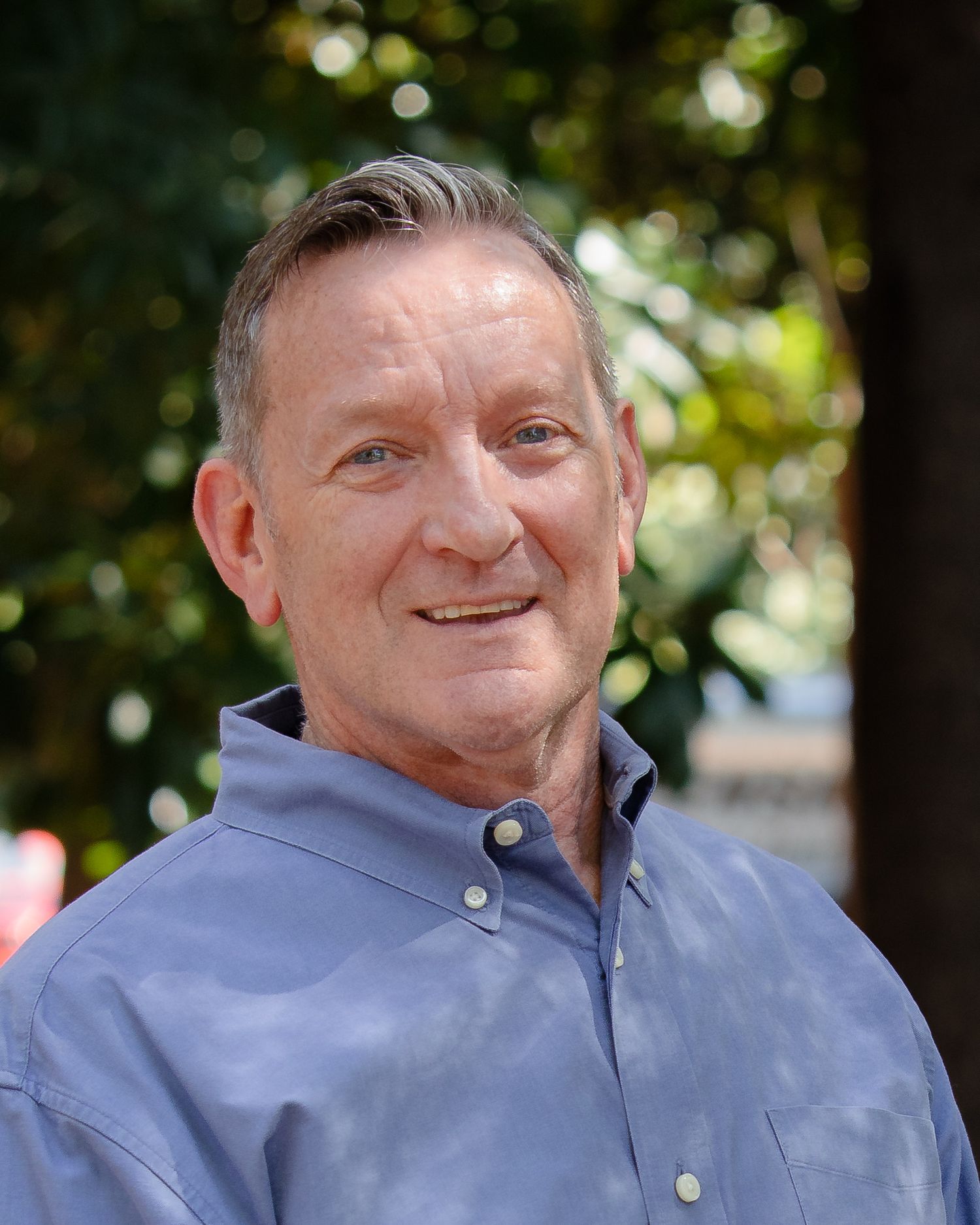 A person wearing a light blue button-up shirt smiles warmly while posing outdoors with trees in the background.