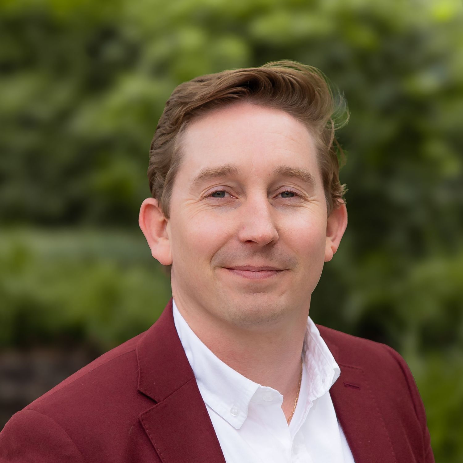 A person wearing a burgundy suit jacket and white shirt smiles warmly at the camera in an outdoor setting.
