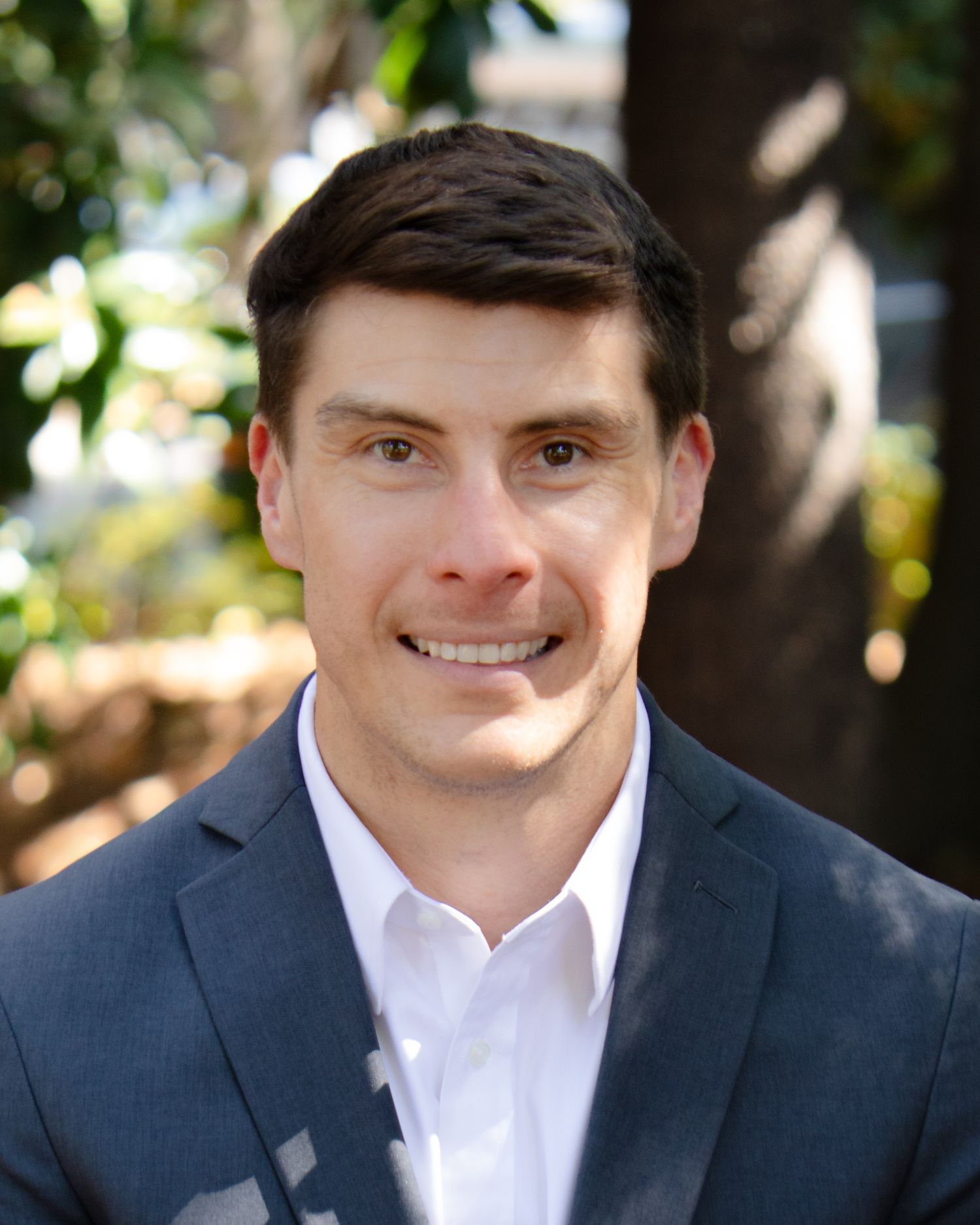 Professional headshot of someone wearing a navy suit jacket and white dress shirt against a blurred outdoor background.
