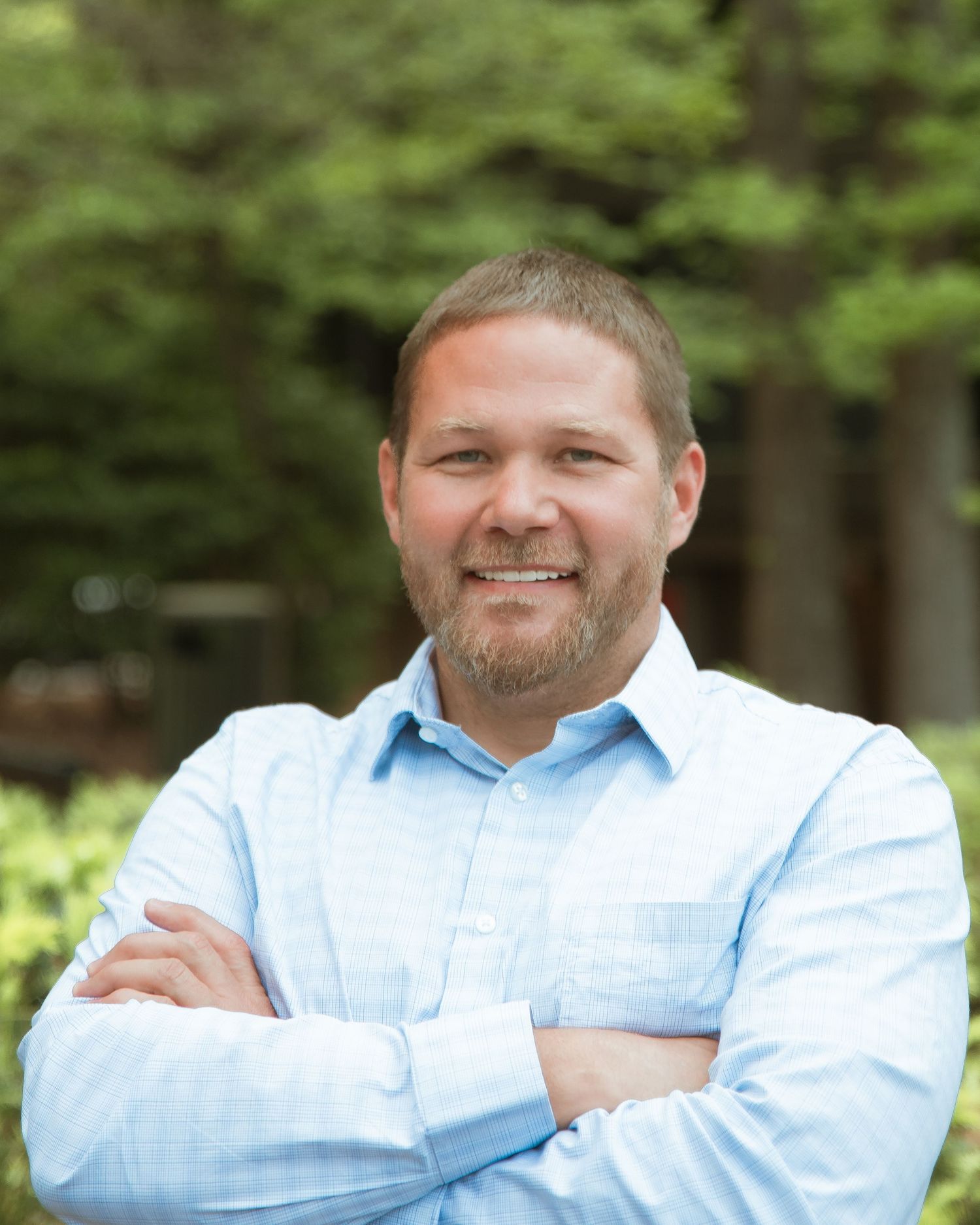 Smiling man with beard, arms crossed, wearing light blue shirt, standing outdoors with green trees in background.