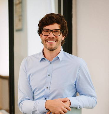 Smiling young professional man with curly hair and glasses wearing a light blue shirt in a bright office.
