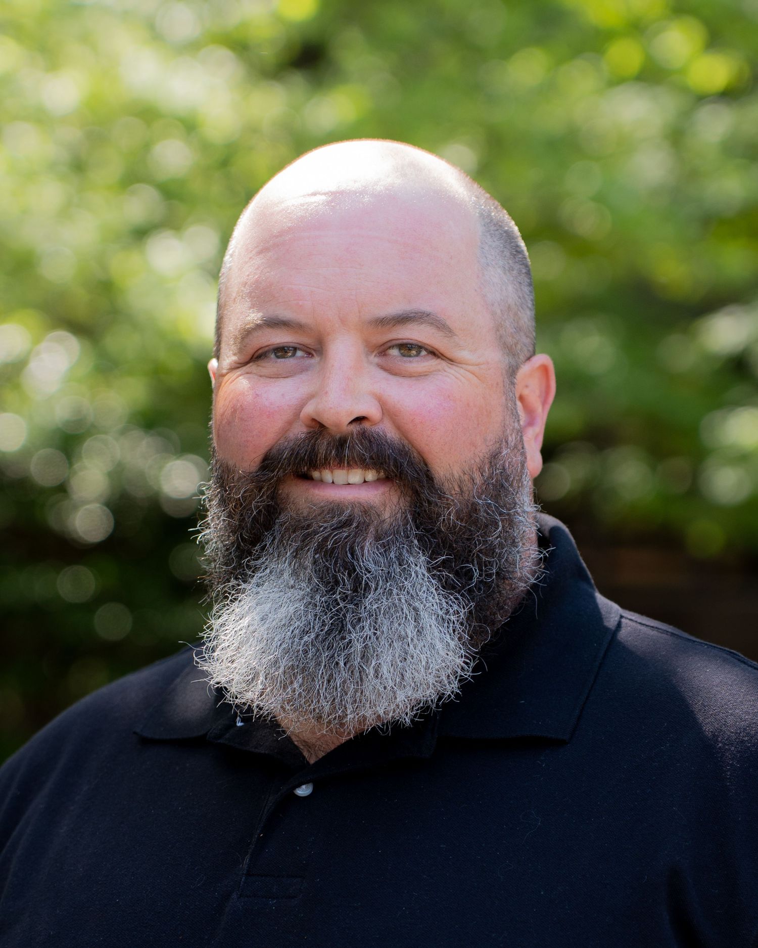 Smiling bald man with a full gray beard wearing a black polo shirt outdoors with green foliage background.