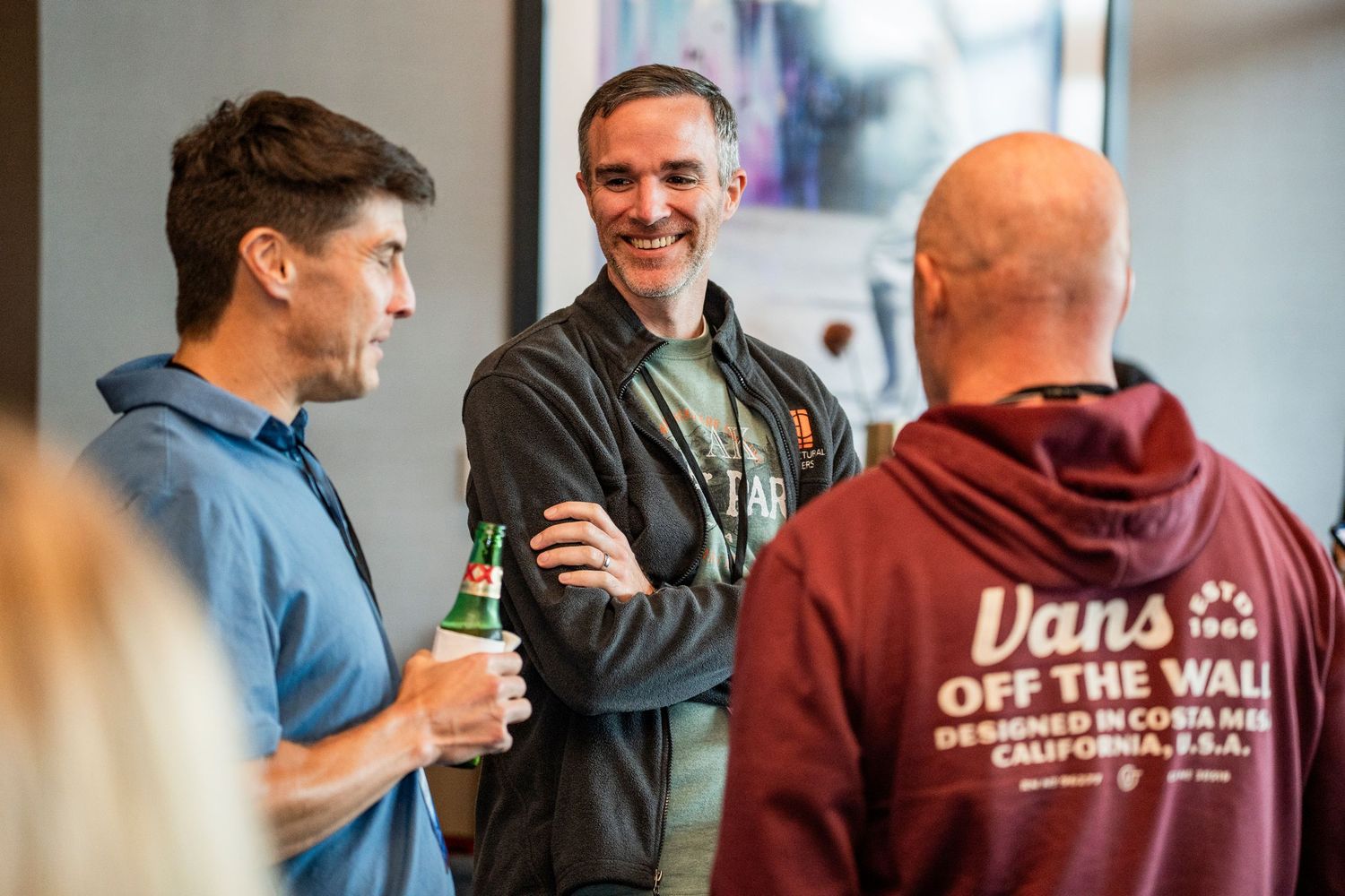 Three people engaged in casual conversation at an indoor gathering, with one wearing a maroon Vans hoodie.