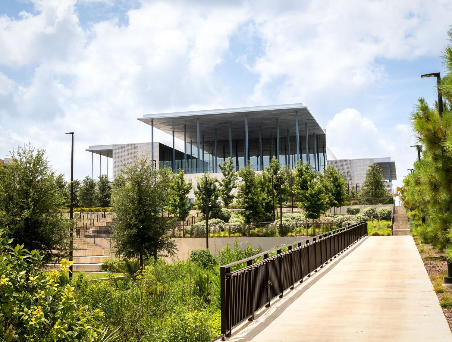 Modern glass building surrounded by lush greenery and a walkway under a partly cloudy sky.