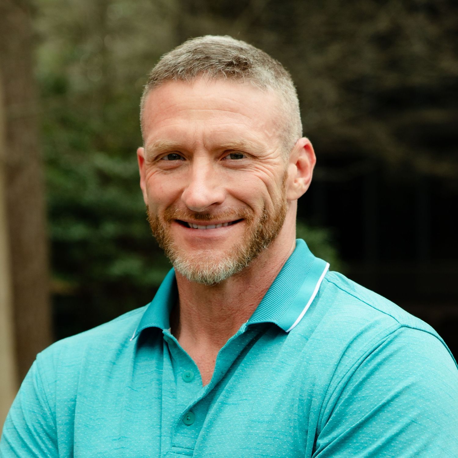 Person wearing turquoise polo shirt smiling warmly in outdoor setting with trees in background.