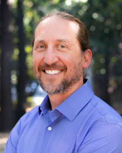 A person in a blue button-down shirt smiles warmly while standing outdoors with trees in the background.