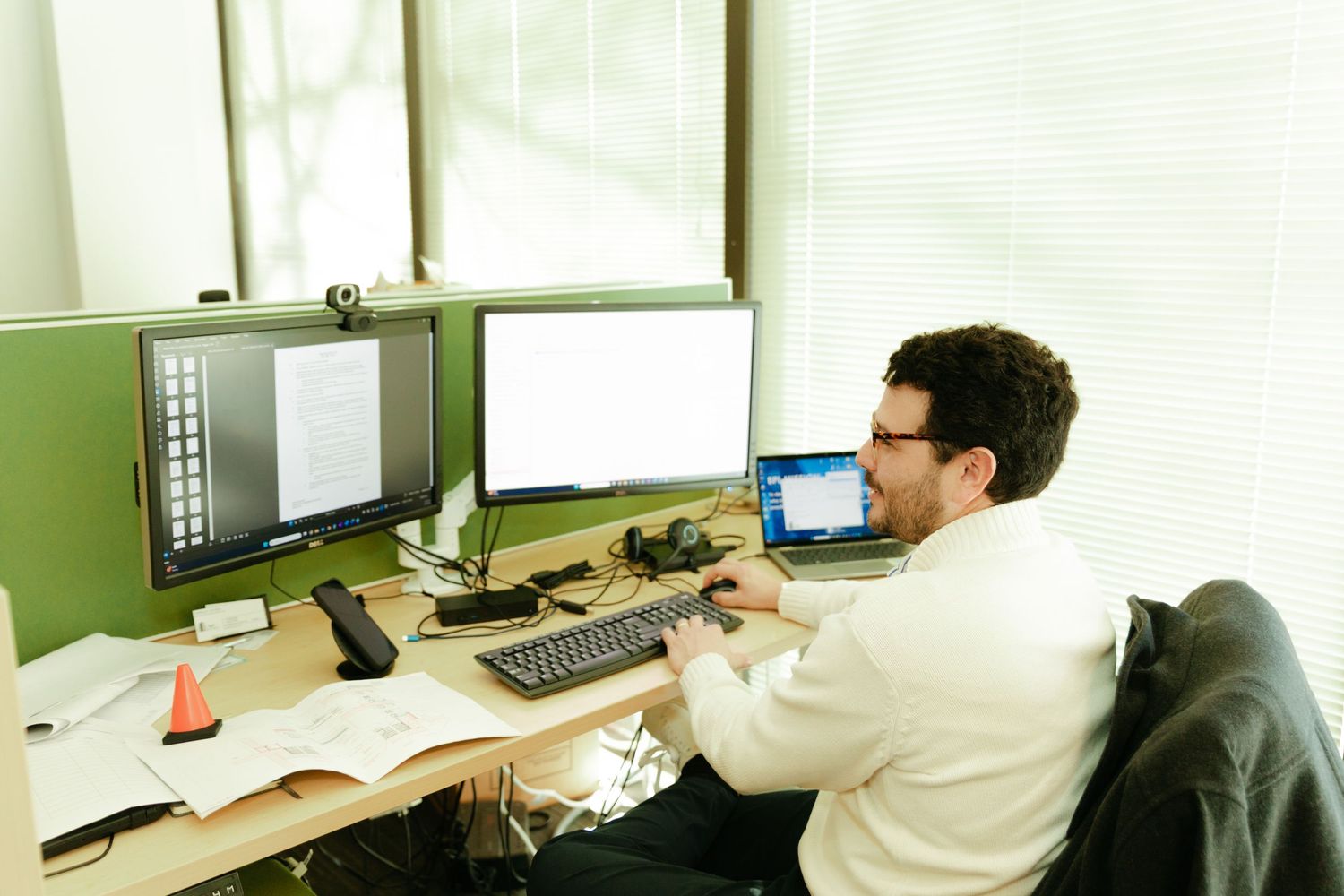 Professional man in glasses working at a multi-monitor desk setup in a modern office cubicle environment.