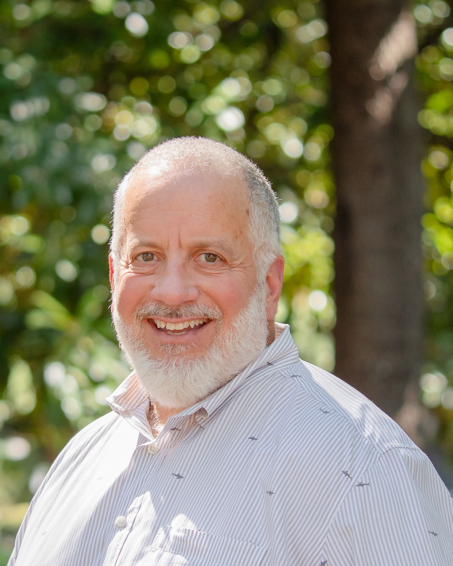 A person with a grey beard wearing a light colored shirt smiles warmly against a blurred green outdoor background.