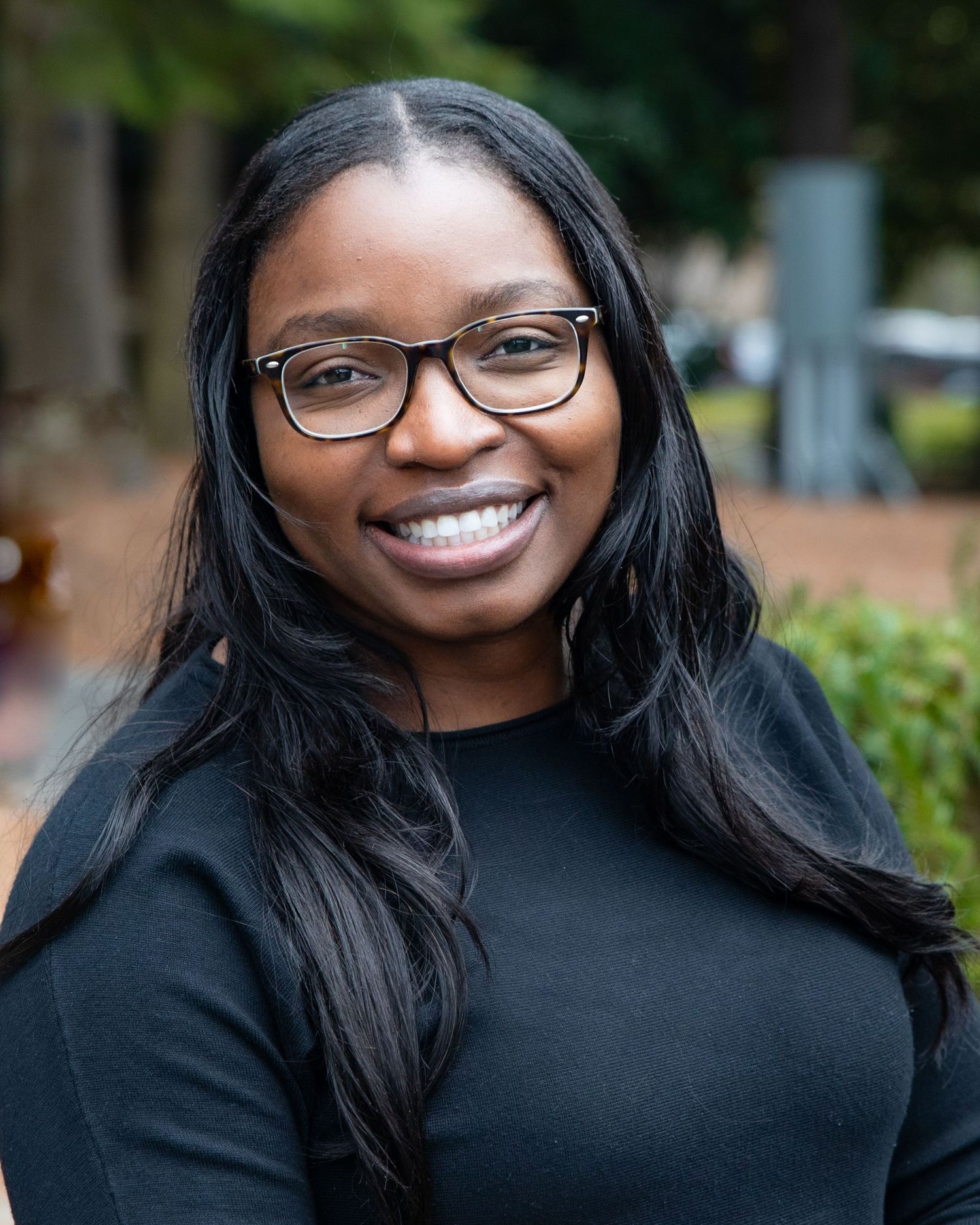 Professional headshot of someone wearing glasses and a black top, smiling warmly against a blurred outdoor background.