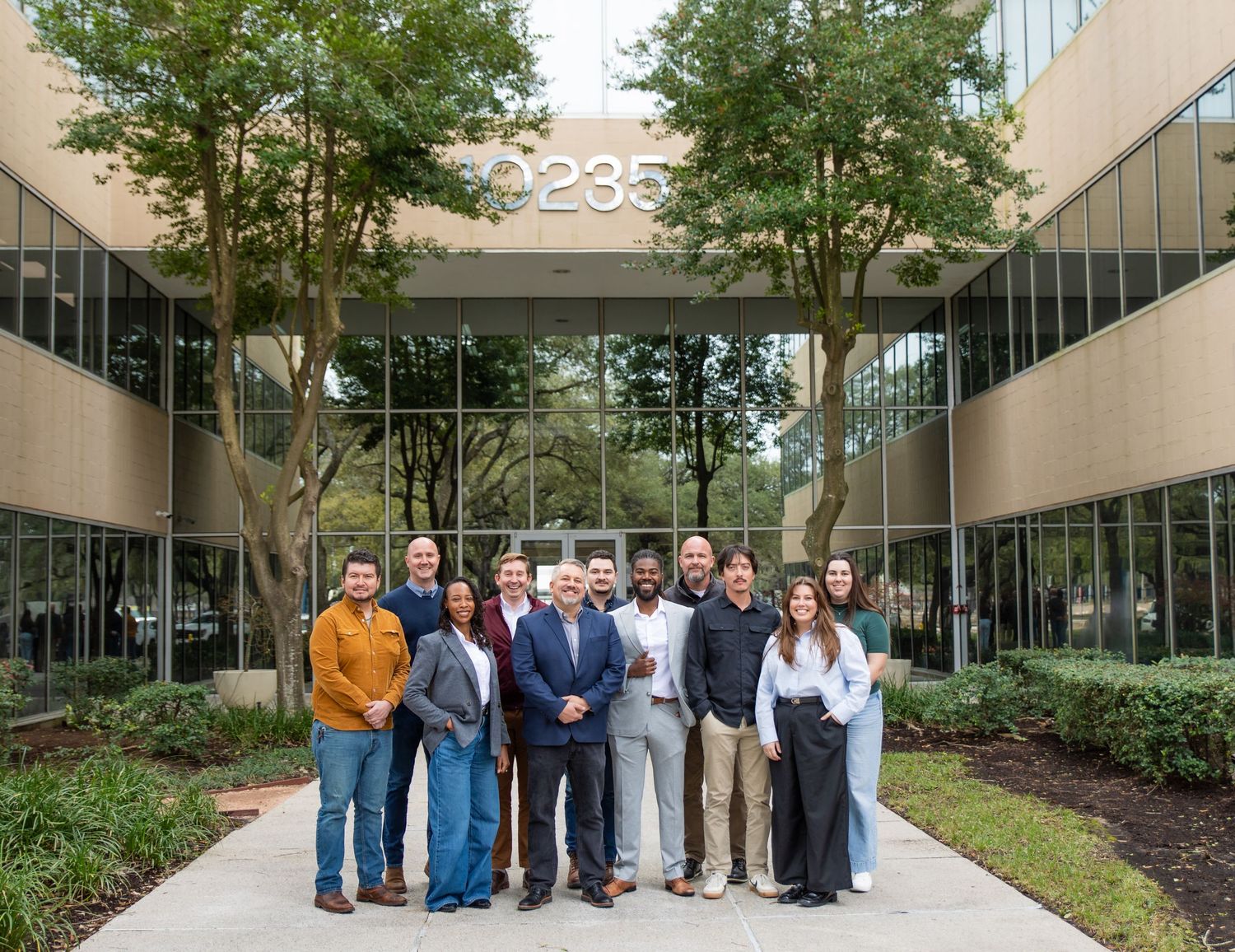 A professional team of employees posing outside a modern office building at 2235, surrounded by trees.
