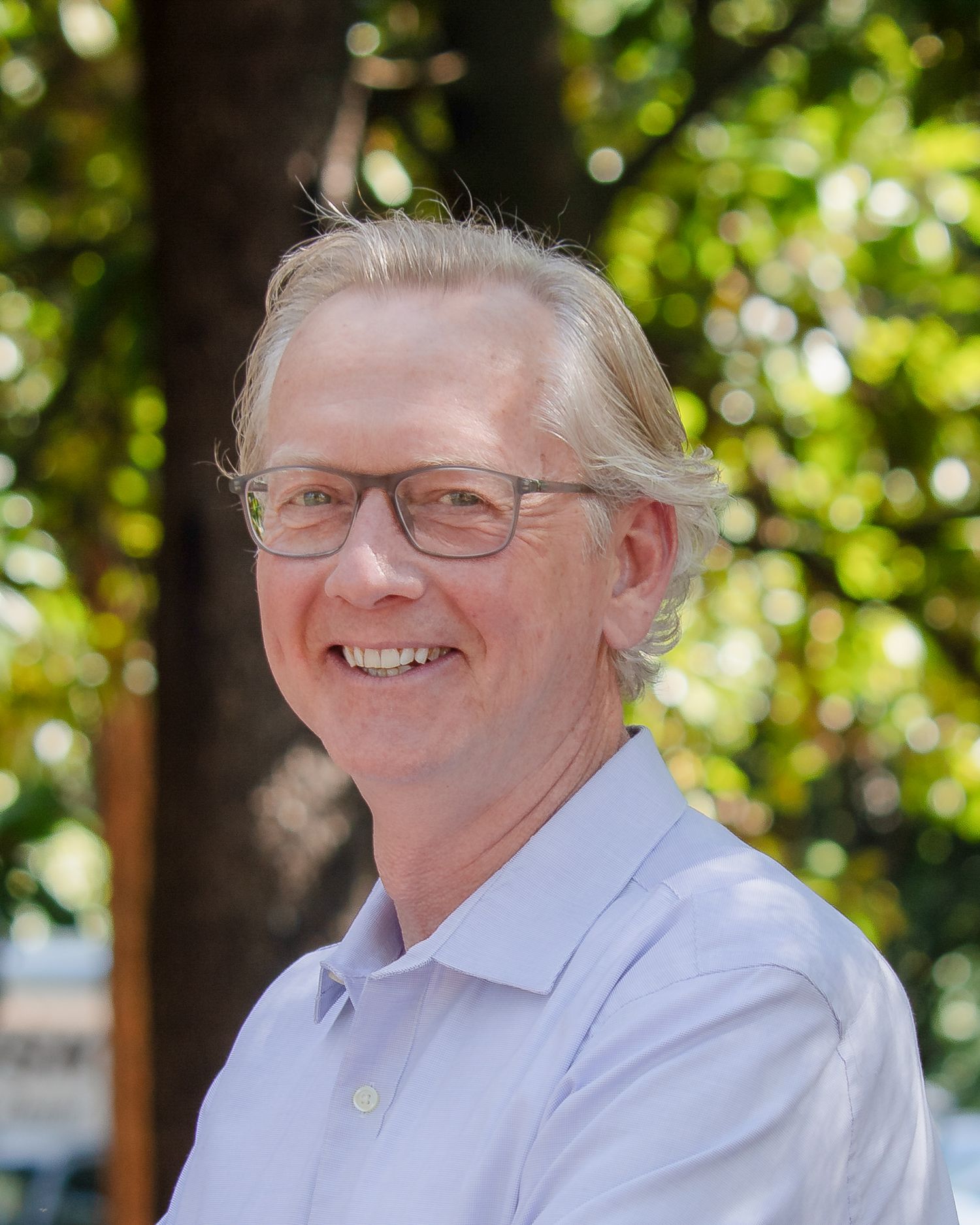 Person wearing glasses and a light colored dress shirt smiles warmly against a green leafy background.