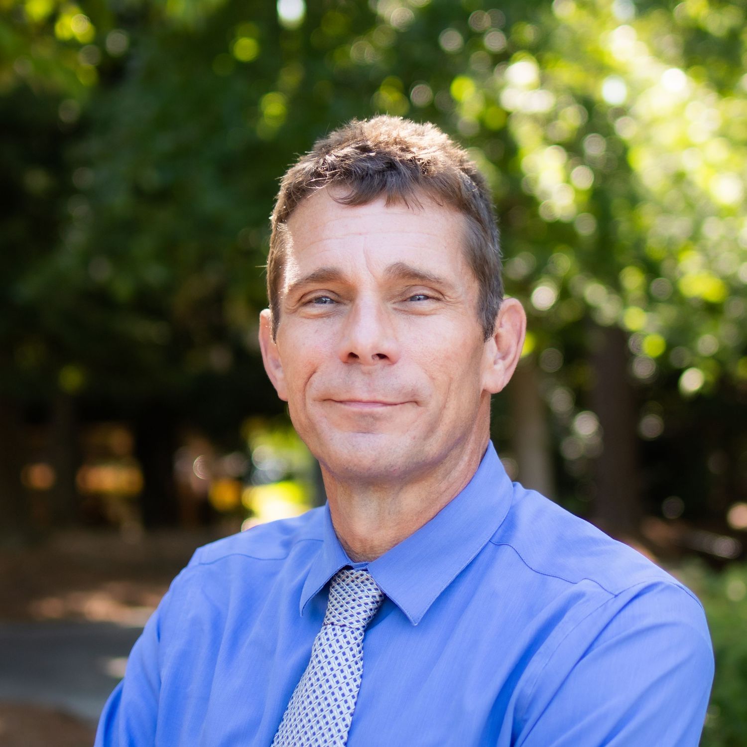 Professional wearing a blue dress shirt and tie smiles warmly against a blurred outdoor background.