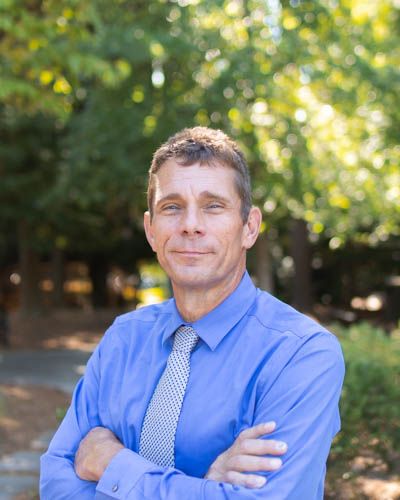 Professional wearing a blue dress shirt and striped tie poses outdoors with crossed arms and friendly expression.