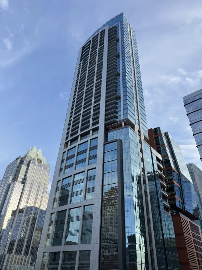 Modern glass skyscraper towers over downtown city skyline against a blue sky with surrounding high-rise buildings.