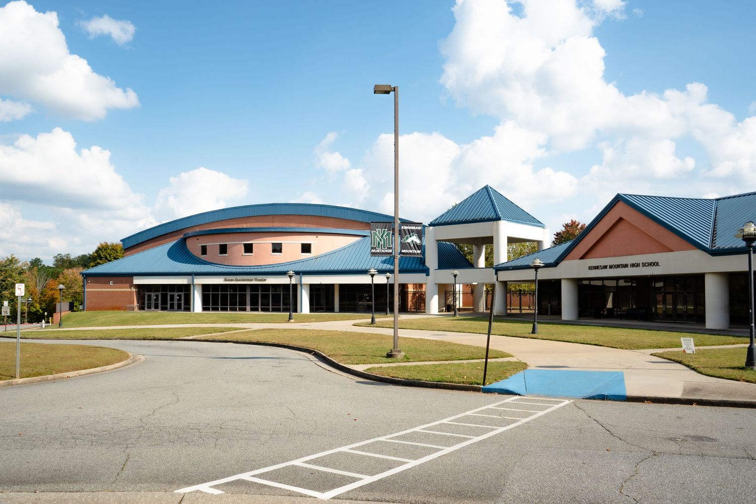 Modern school building exterior with curved blue roof and brick walls under sunny blue sky with white clouds.