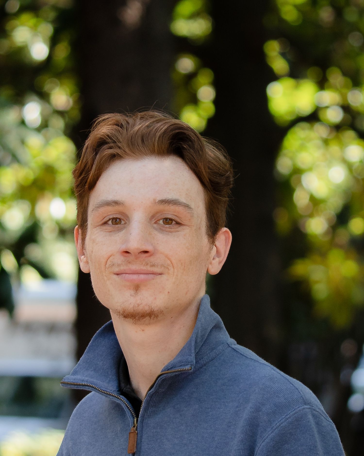 A person in a blue zip-up jacket smiles playfully at the camera in an outdoor setting with bokeh lighting effects.