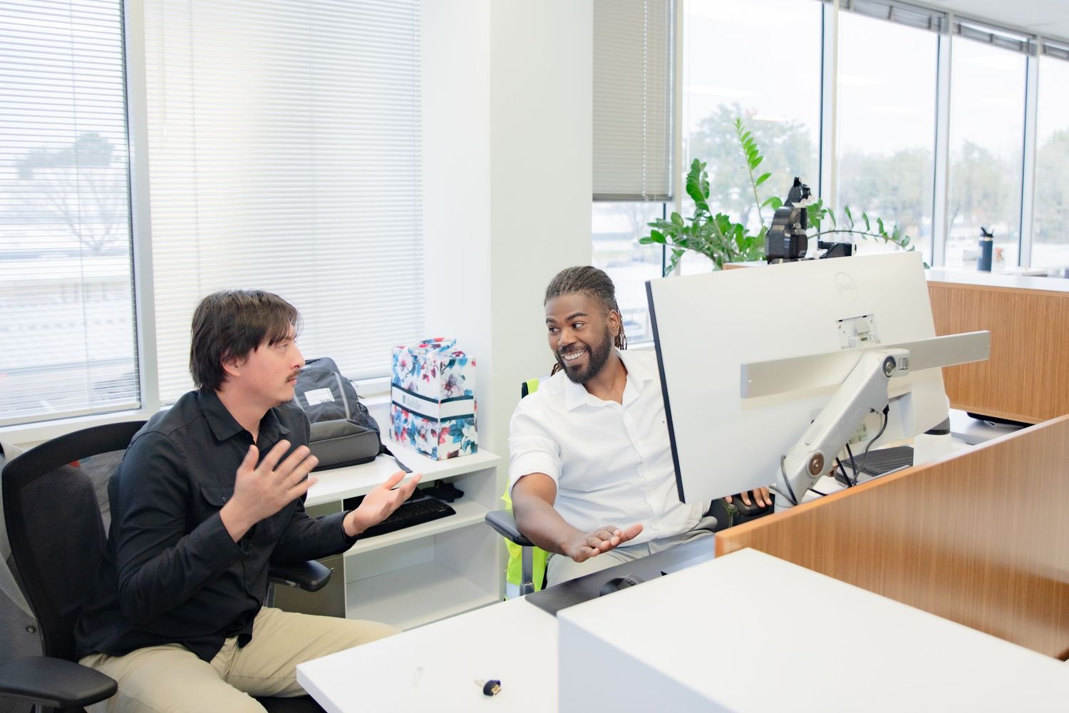 Two colleagues smiling and conversing at a modern office reception desk with plants and computer monitors.